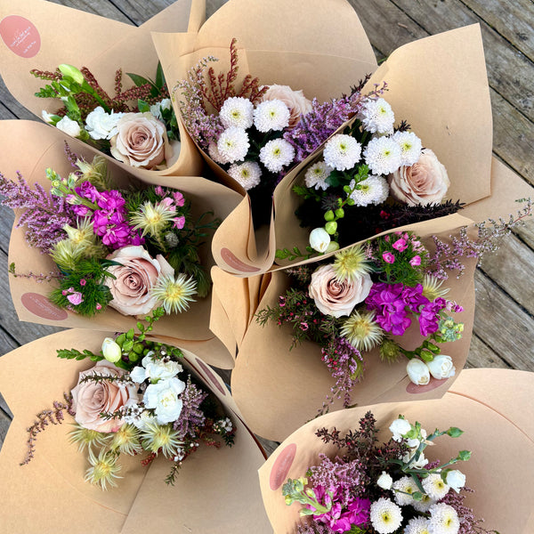 Five floral bouquets wrapped in brown kraft paper, arranged on a weathered wooden deck. The bouquets feature light pink roses, white daisies, purple filler flowers, and green foliage.
